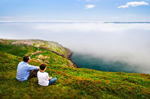 A man and boy looking out across a foggy sea towards the horizon from a green headland in Newfoundland, Canada